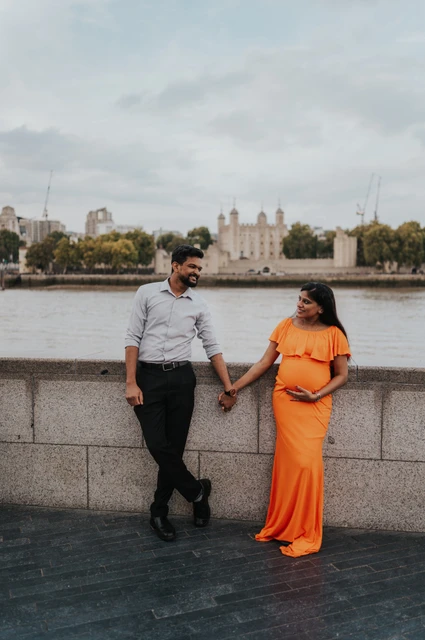 London maternity photoshoot: hand in hand across from the Tower of London. A couple hold hands beside the Queen’s Walk river wall, with the woman in an orange dress and the Tower of London across the Thames during their London maternity photoshoot.
