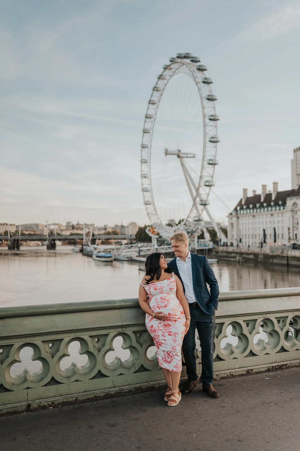 The couple looking at one another by the Westminster Bridge balustrade with the London Eye behind during their London maternity photoshoot.