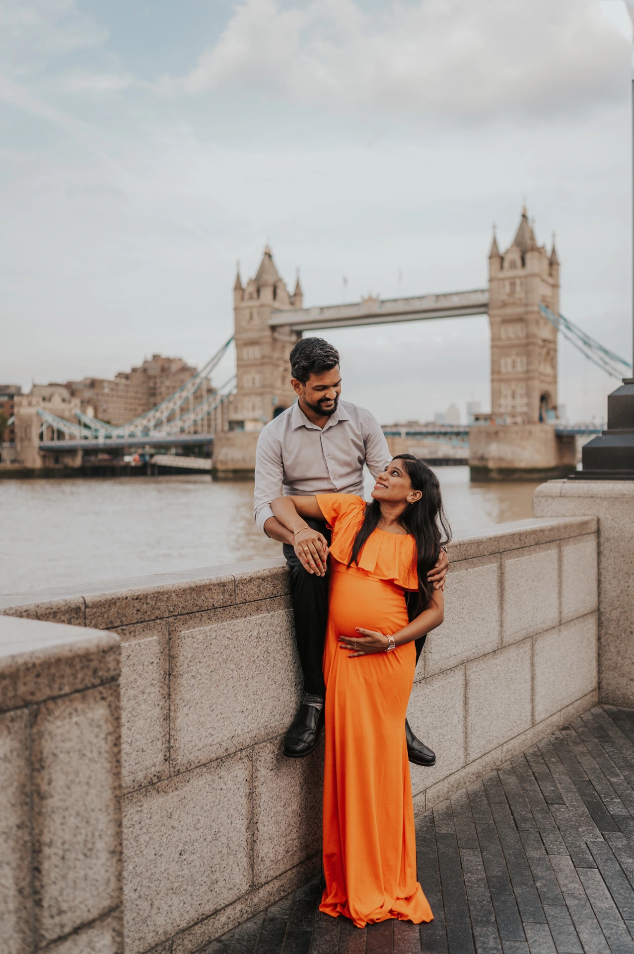 A pregnant woman in an orange dress looks up towards her partner as he sits on the Queen’s Walk river wall, with Tower Bridge behind during their London maternity photoshoot.