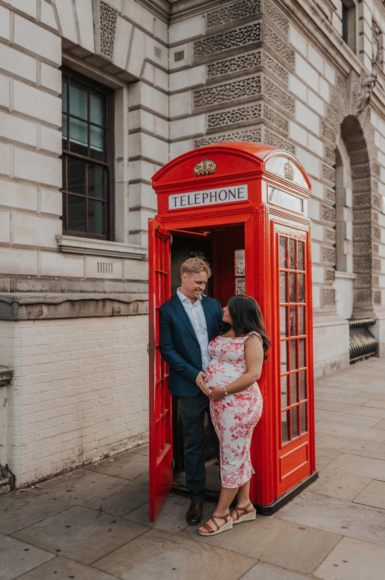 Full-length portrait of the couple together in the open doorway of a red telephone box during their London maternity photoshoot.