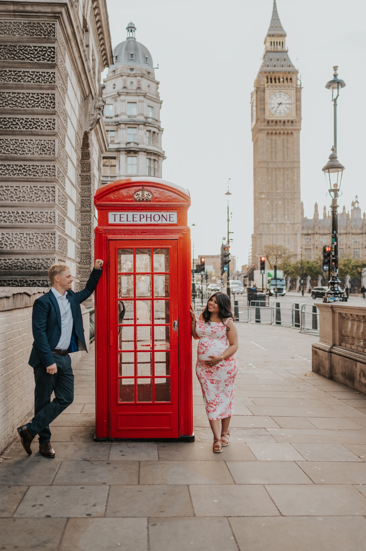 The expectant couple standing on opposite sides of a red telephone box, with Big Ben behind, during their London maternity photoshoot.