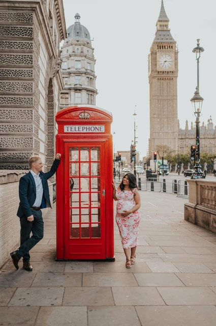 A playful telephone box portrait at a London maternity photoshoot The expectant couple standing on opposite sides of a red telephone box, with Big Ben behind, during their London maternity photoshoot.