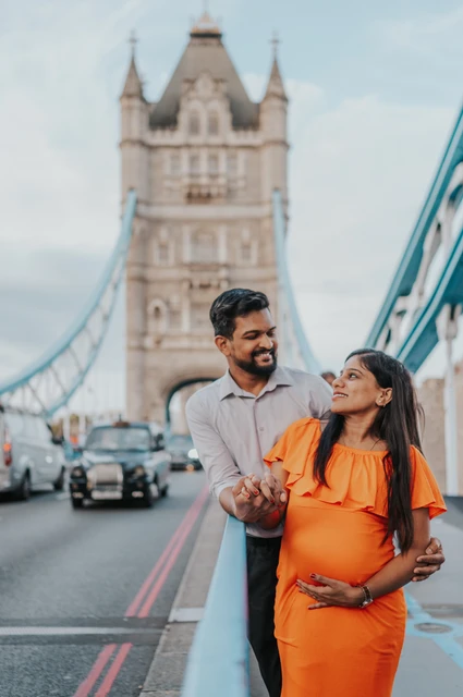 A close couple portrait shows a pregnant woman in an orange dress looking towards her partner beside the blue Tower Bridge railing, with a bridge tower behind during their London maternity photoshoot.