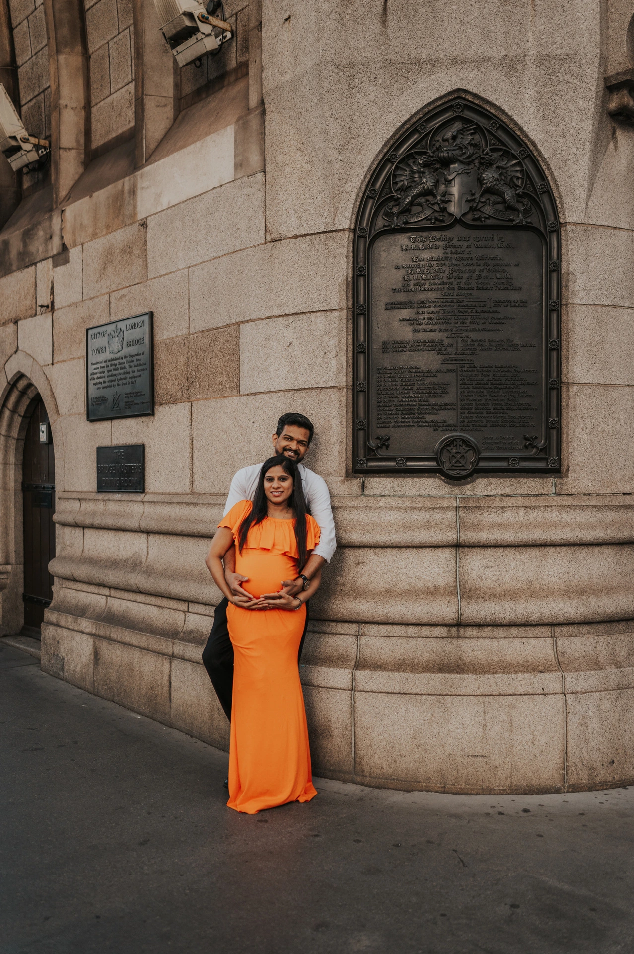 A couple face the camera beside the curved Tower Bridge stonework and decorative plaque, with the woman holding her bump in an orange dress during their London maternity photoshoot.