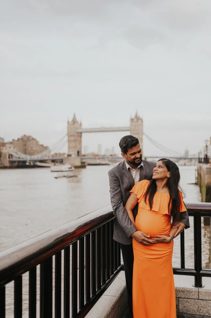 London maternity photoshoot: together at the riverside railing. A couple stand together at a dark riverside railing, with her orange dress in the foreground and Tower Bridge across the Thames during their London maternity photoshoot.