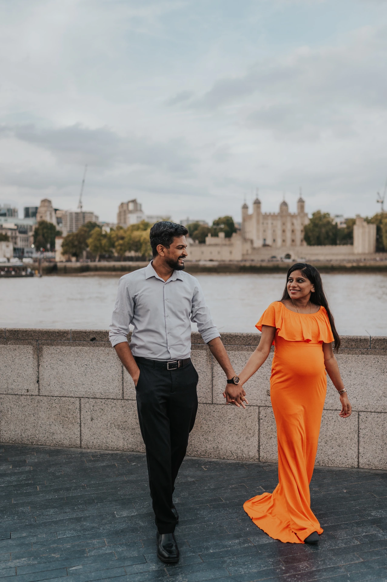 A couple walk hand in hand on the Queen’s Walk, smiling towards each other with the orange dress moving beside the river wall during their London maternity photoshoot.