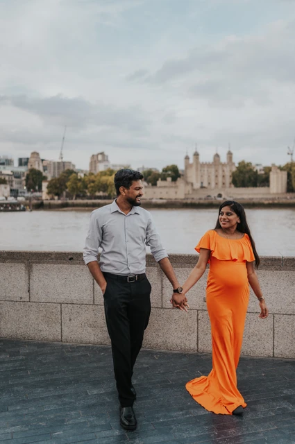 London maternity photoshoot: an orange-dress walk along the river. A couple walk hand in hand on the Queen’s Walk, smiling towards each other with the orange dress moving beside the river wall during their London maternity photoshoot.