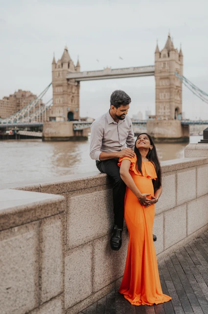 London maternity photoshoot: a couple portrait beside the stone river wall. A man sits on the Queen’s Walk river wall behind his pregnant partner in an orange dress, with Tower Bridge across the water during their London maternity photoshoot.