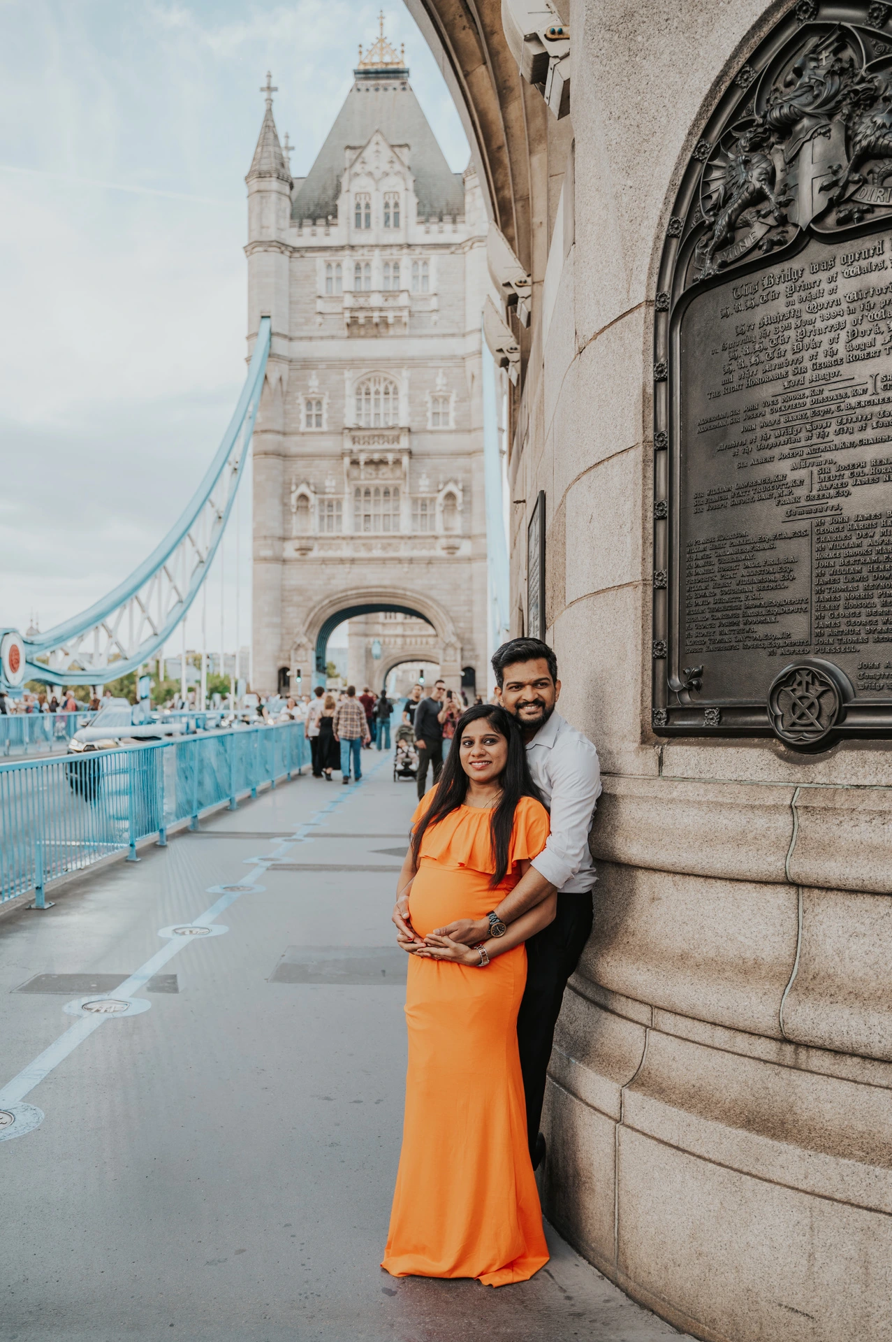 A couple stand together beside Tower Bridge’s stone wall and commemorative plaque, with her orange dress and a bridge tower in view during their London maternity photoshoot.