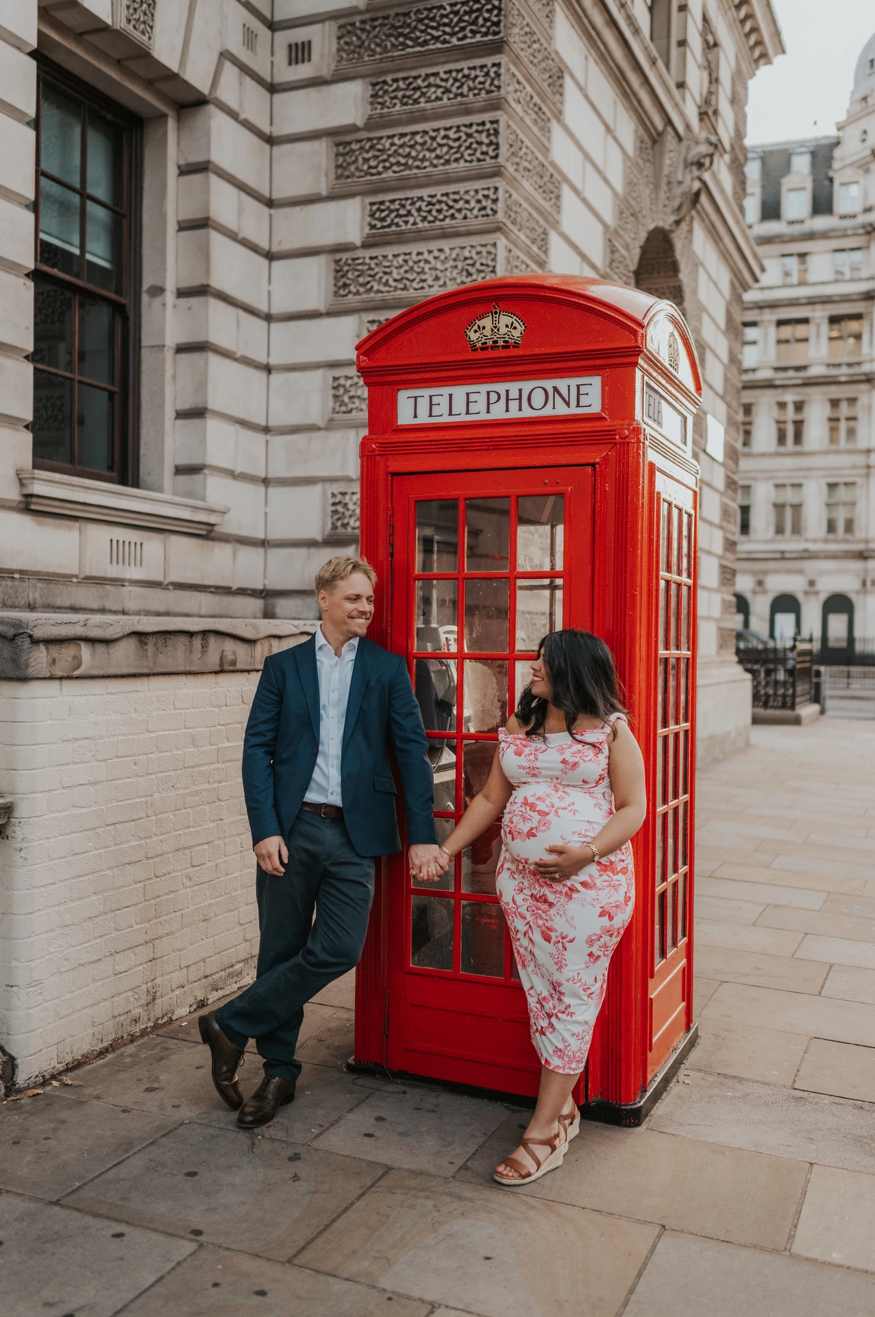 Angled full-length portrait of the couple holding hands on either side of a red telephone box during their London maternity photoshoot.