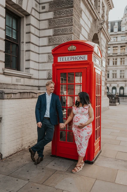 A Westminster street portrait at a London maternity photoshoot Angled full-length portrait of the couple holding hands on either side of a red telephone box during their London maternity photoshoot.