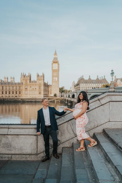 A riverside steps portrait at a London maternity photoshoot The mother-to-be in a floral dress standing a step above her partner beside the Thames during their London maternity photoshoot.