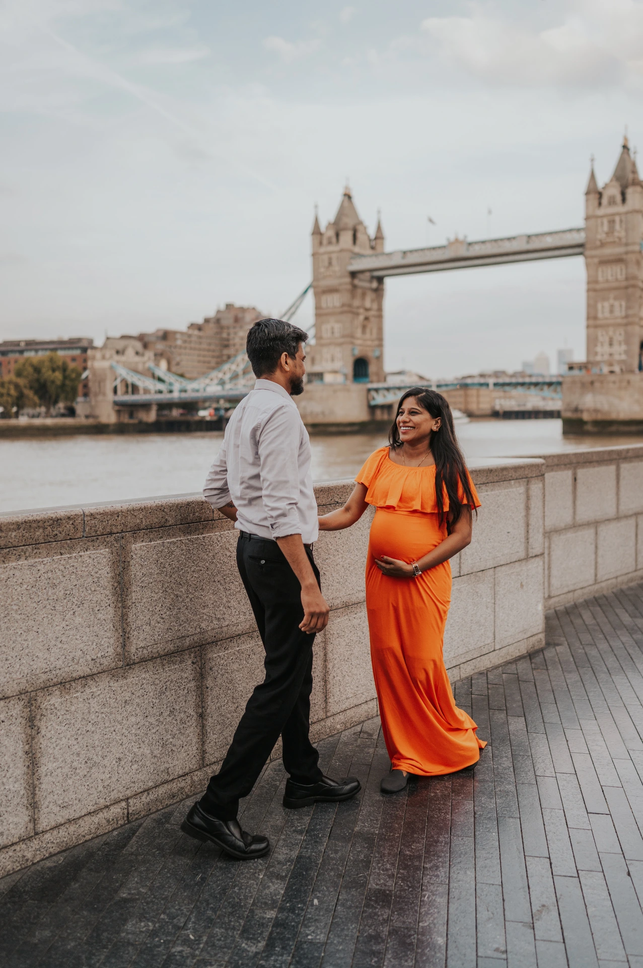 A couple move towards each other beside the Queen’s Walk wall, with the woman in an orange dress and Tower Bridge in the background during their London maternity photoshoot.