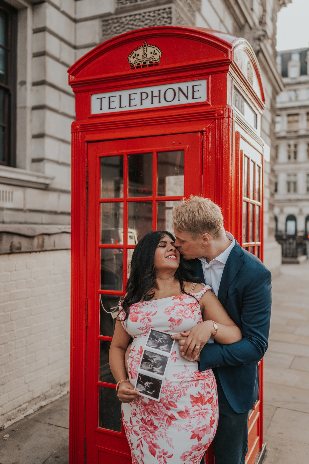 The couple sharing a close moment beside a red telephone box while she holds their ultrasound scan during a London maternity photoshoot.