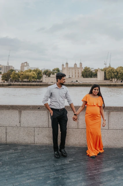 London maternity photoshoot: a hand-in-hand walk beside the Thames. A couple look down as they walk hand in hand beside the Queen’s Walk wall, with her orange dress and the Thames behind during their London maternity photoshoot.
