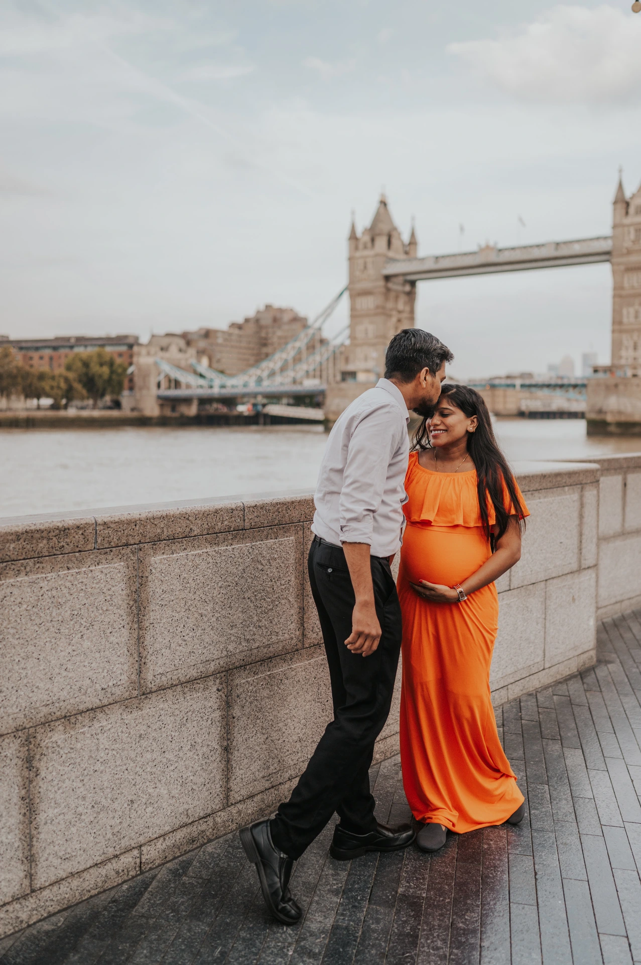 A man leans towards his pregnant partner’s forehead beside the Queen’s Walk river wall, with her orange dress and Tower Bridge in view during their London maternity photoshoot.