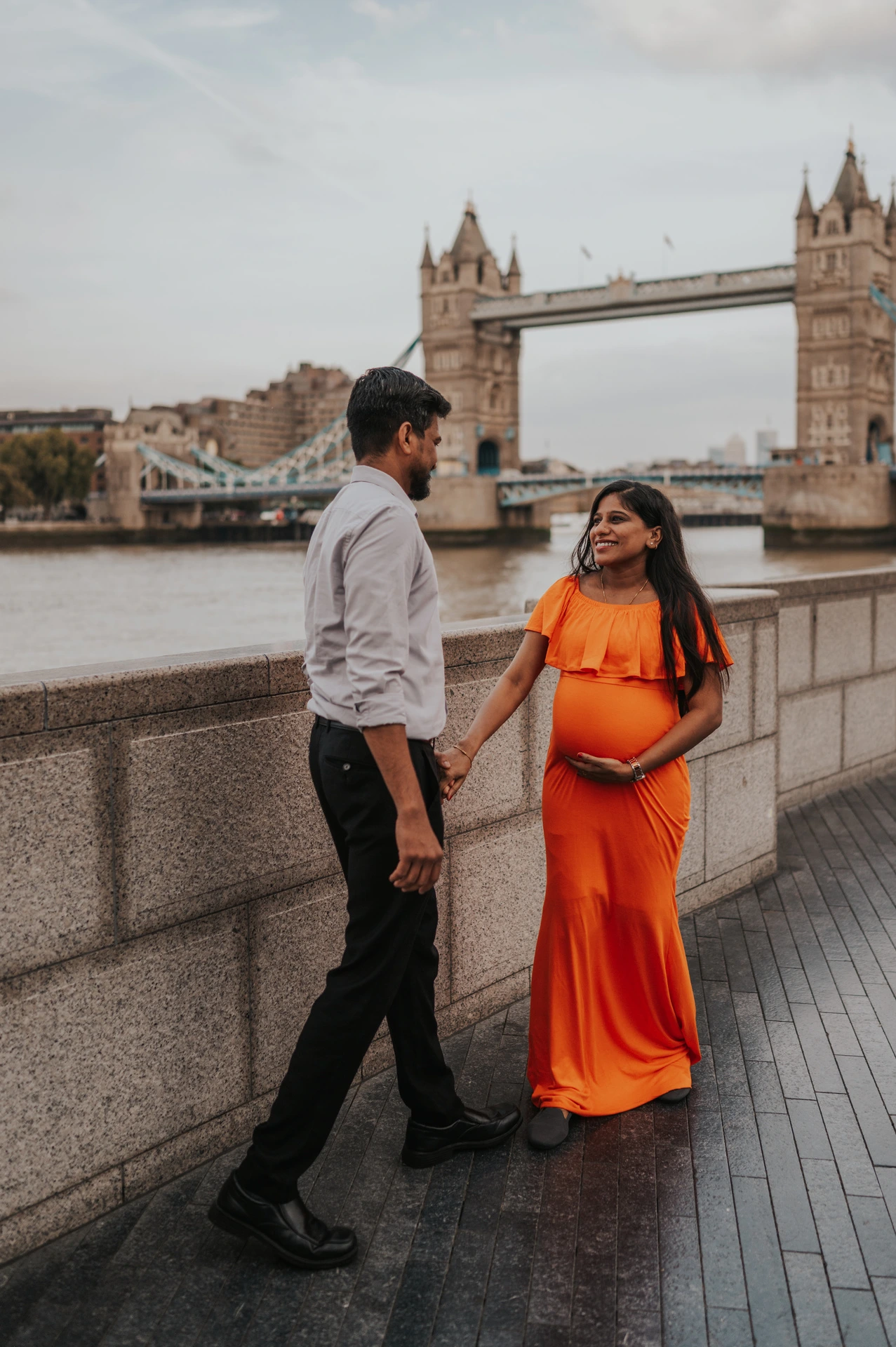 A full-length portrait shows a couple facing each other beside the Queen’s Walk river wall, with the woman’s orange dress and Tower Bridge behind during their London maternity photoshoot.