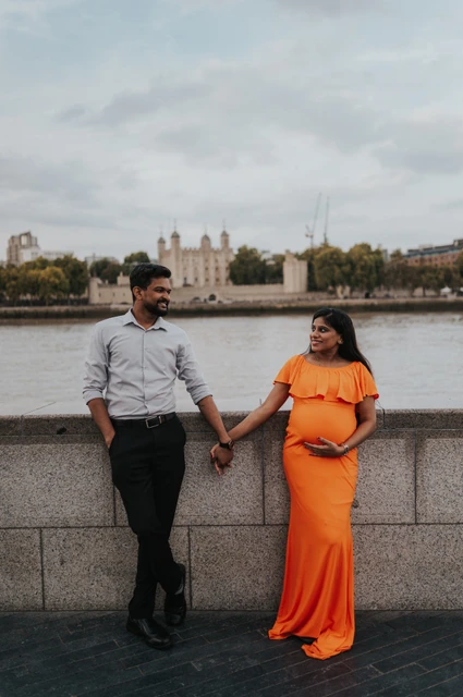 London maternity photoshoot: full-length couple portrait across from the Tower. A full-length portrait shows a couple holding hands beside the Queen’s Walk wall, with her long orange dress and the Tower of London across the river during their London maternity photoshoot.