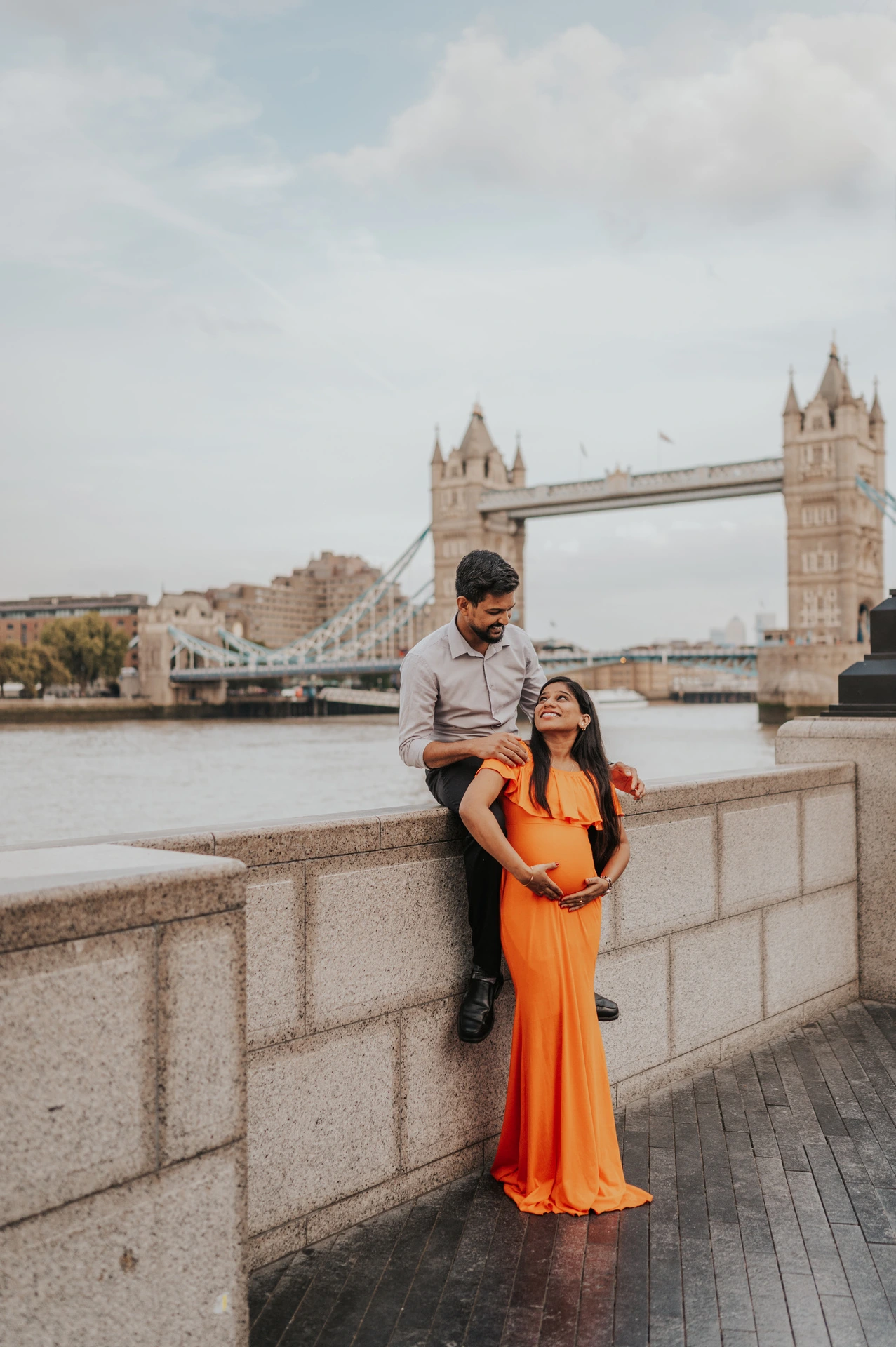 London maternity photoshoot: full-length portrait with Tower Bridge across the Thames. A full-length portrait shows a pregnant woman in an orange dress with her partner seated on the river wall behind her, beneath the view of Tower Bridge during their London maternity photoshoot.