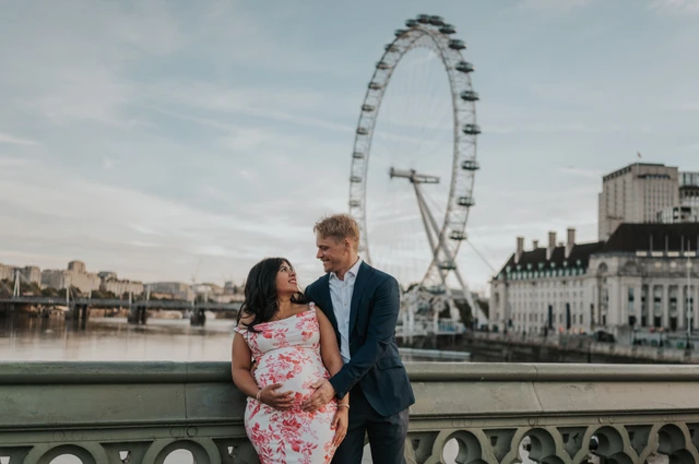 Landscape portrait of the couple holding her bump beside Westminster Bridge railing with the London Eye behind during their London maternity photoshoot.