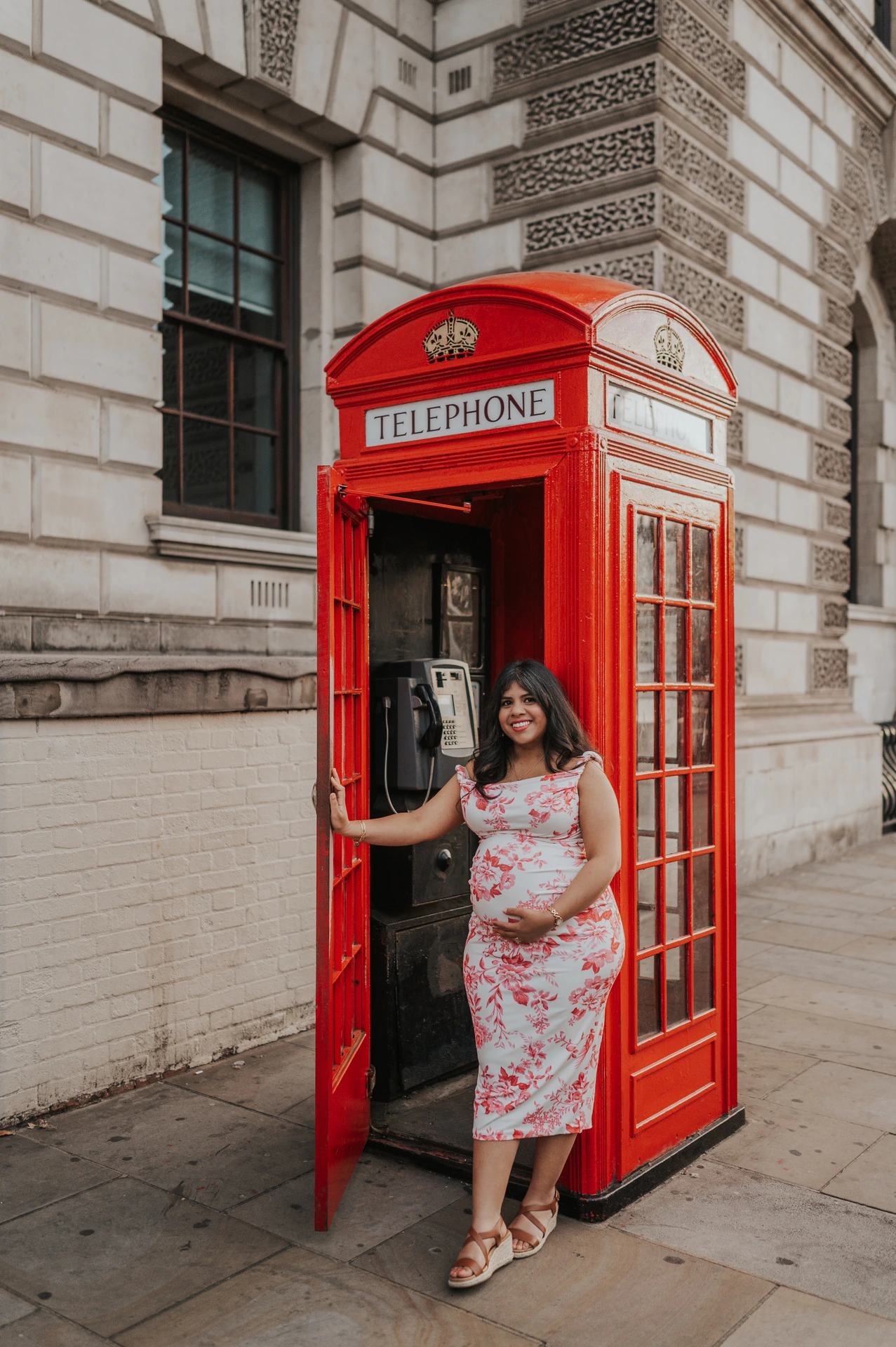The mother-to-be standing at the open door of a red telephone box against a carved stone facade during her London maternity photoshoot.