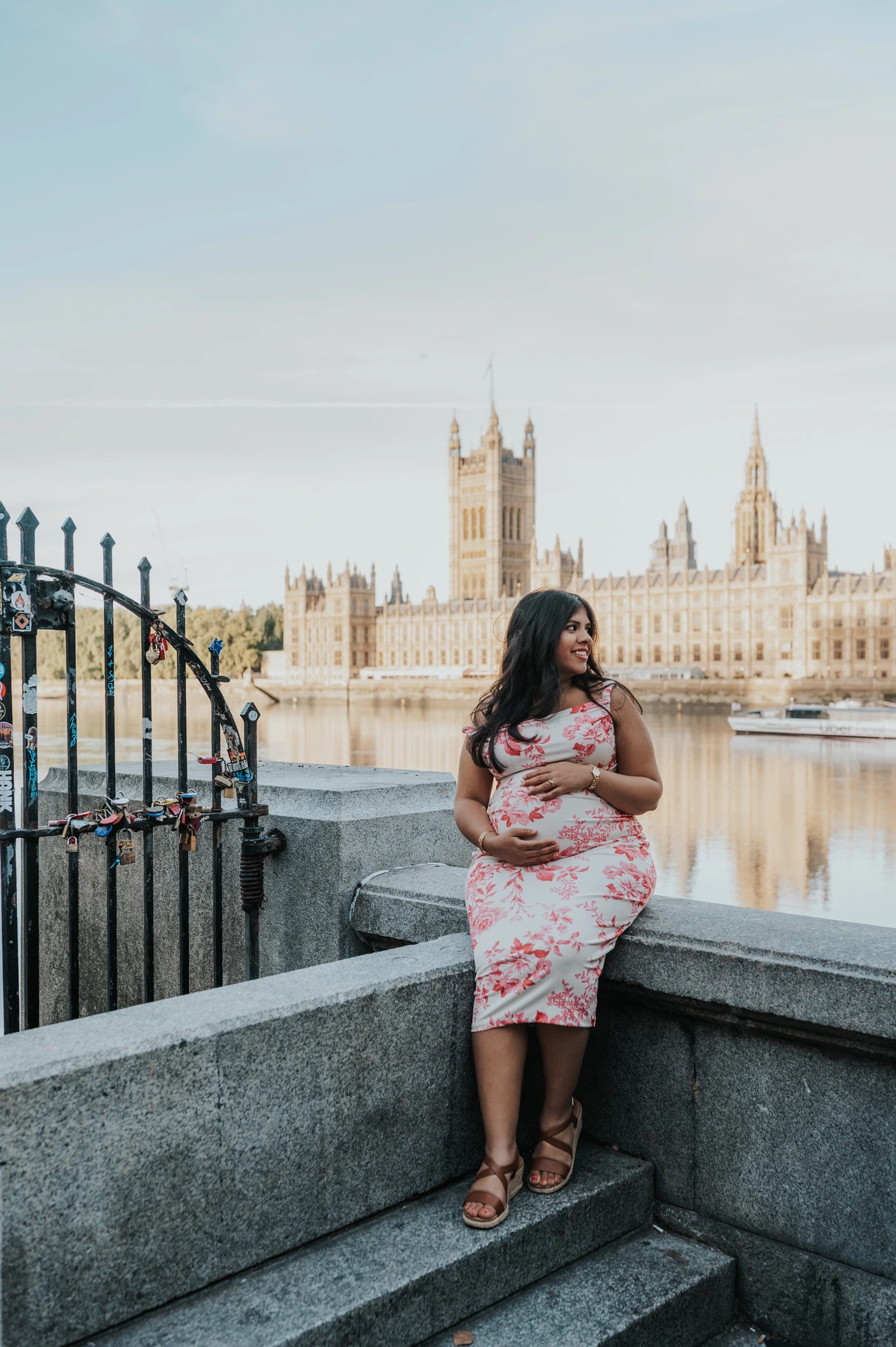 The mother-to-be standing by a riverside stone wall with the Houses of Parliament across the Thames during her London maternity photoshoot.