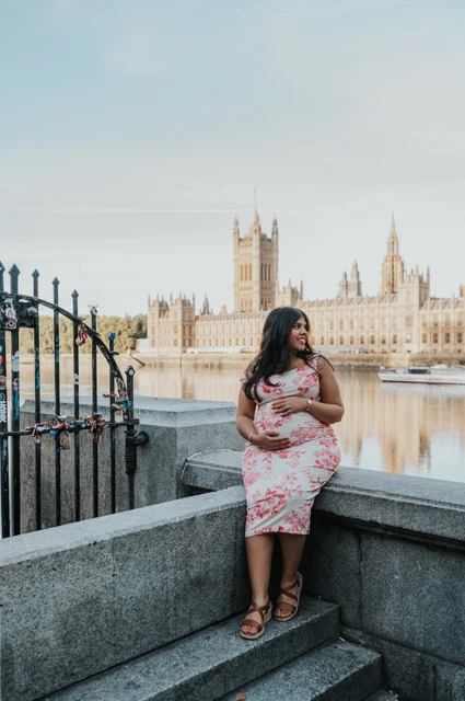 The mother-to-be standing by a riverside stone wall with the Houses of Parliament across the Thames during her London maternity photoshoot.