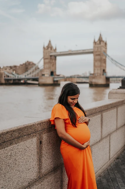 London maternity photoshoot: looking down at her bump by the river. A pregnant woman in an orange dress looks down at her bump beside the Queen’s Walk river wall, with Tower Bridge beyond during their London maternity photoshoot.