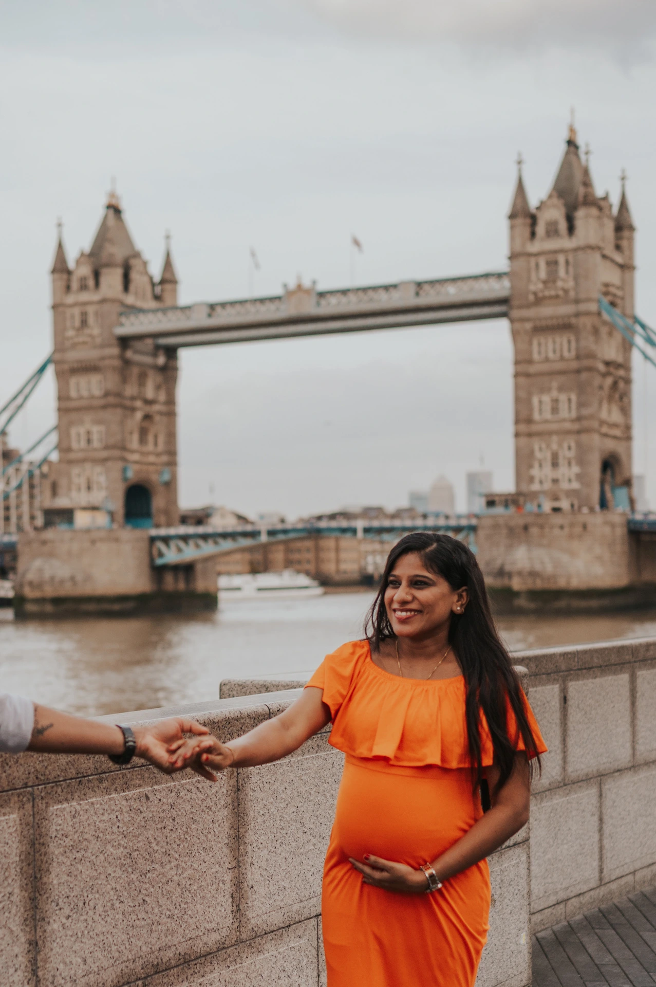 A close portrait shows a pregnant woman in an orange dress holding her partner’s hand at the edge of the frame, with Tower Bridge behind during their London maternity photoshoot.