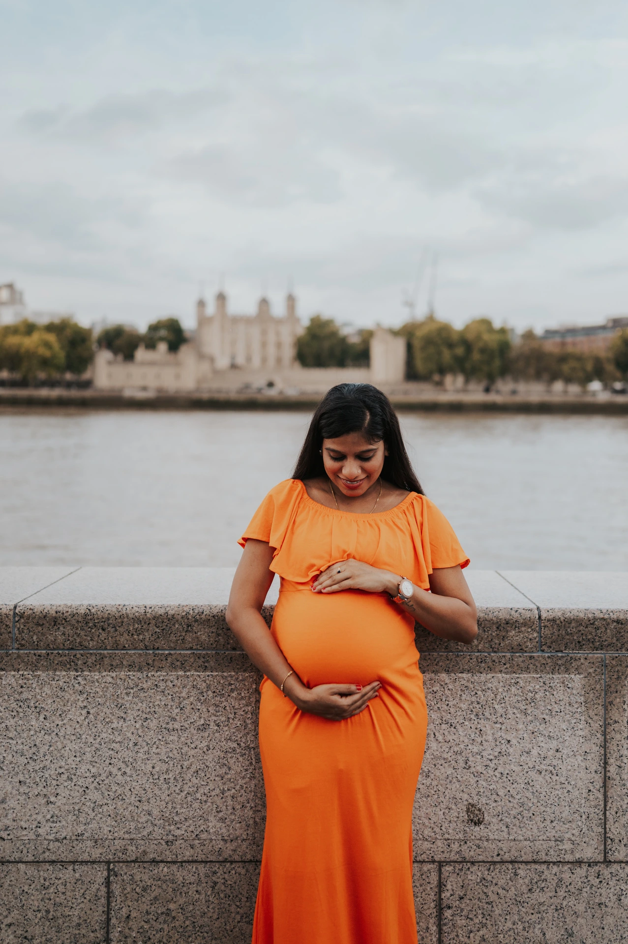 A pregnant woman looks down at her bump in an orange dress beside the Queen’s Walk wall, with the Thames and Tower of London beyond during their London maternity photoshoot.