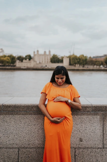 London maternity photoshoot: a quiet orange-dress portrait by the river wall. A pregnant woman looks down at her bump in an orange dress beside the Queen’s Walk wall, with the Thames and Tower of London beyond during their London maternity photoshoot.
