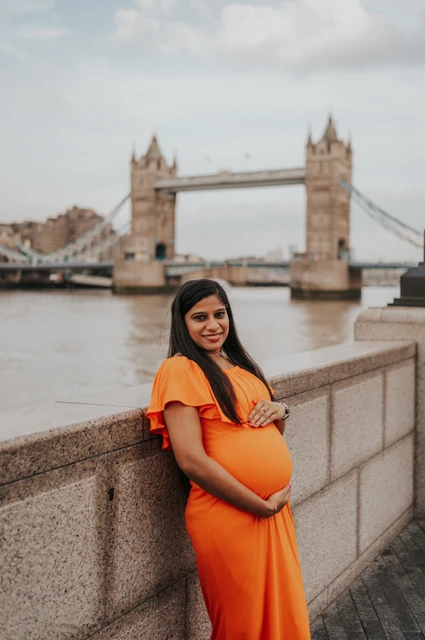 London maternity photoshoot: an orange-dress portrait with Tower Bridge. A pregnant woman in an orange dress faces the camera beside the Queen’s Walk stone wall, with Tower Bridge behind her during their London maternity photoshoot.