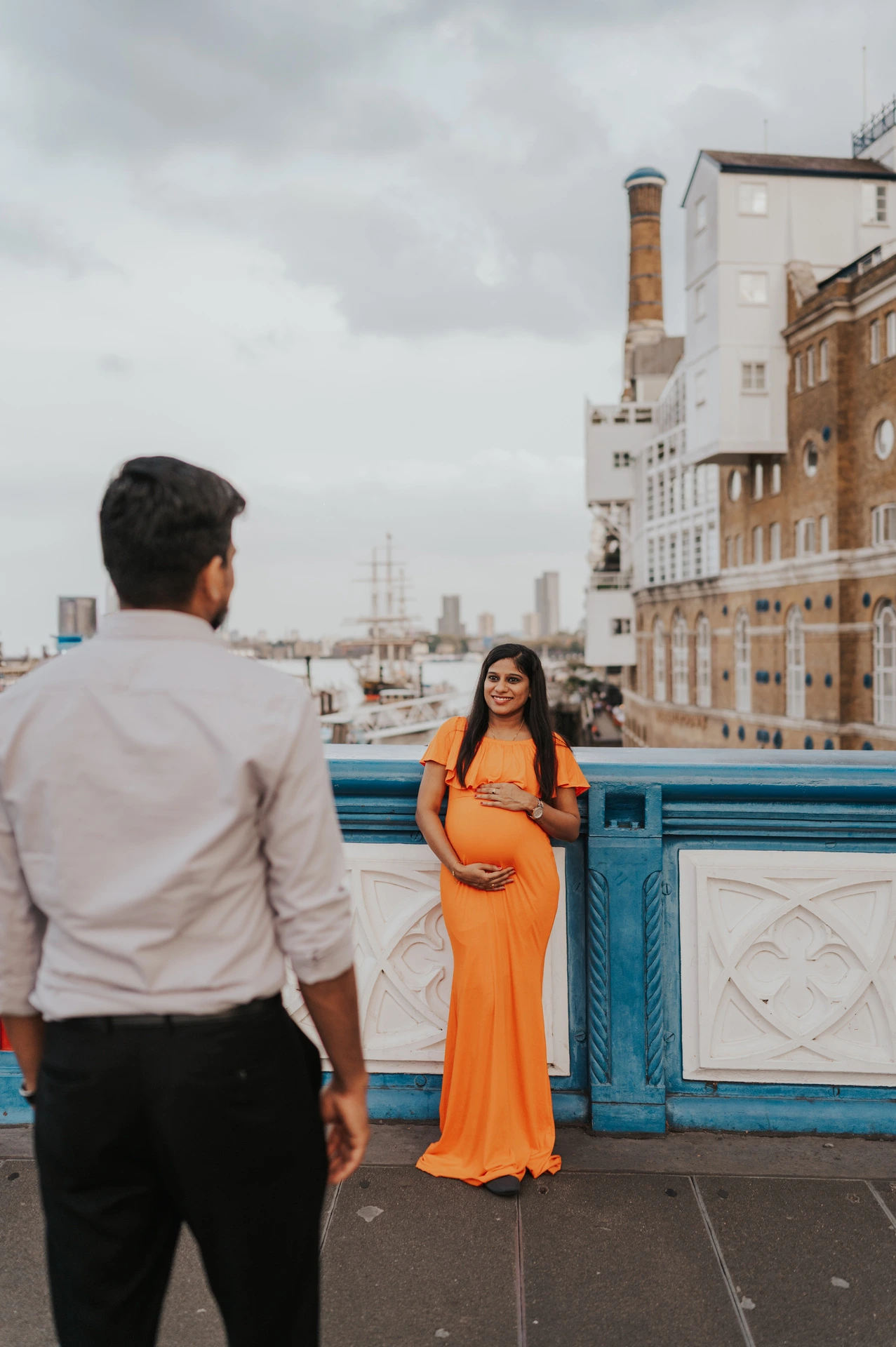 A man faces his pregnant partner in an orange dress beside the blue Tower Bridge railing, with Butler’s Wharf beyond during their London maternity photoshoot.