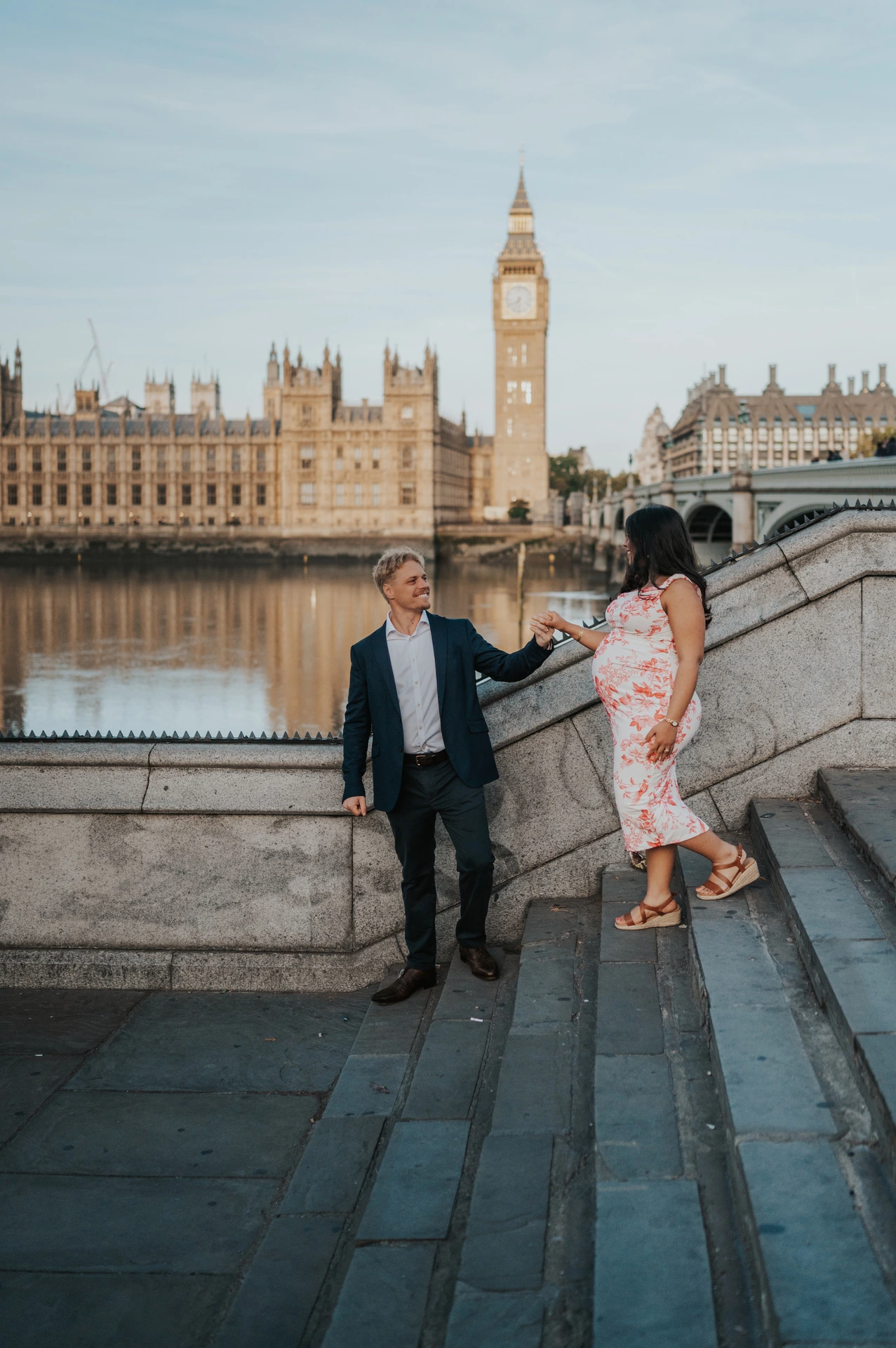 The couple holding hands on stone riverside steps with Big Ben and the Houses of Parliament across the Thames during their London maternity photoshoot.