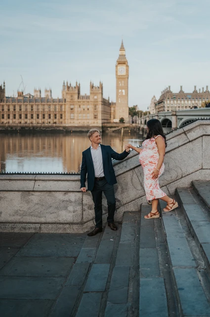 London maternity photoshoot on the riverside steps at Westminster The couple holding hands on stone riverside steps with Big Ben and the Houses of Parliament across the Thames during their London maternity photoshoot.