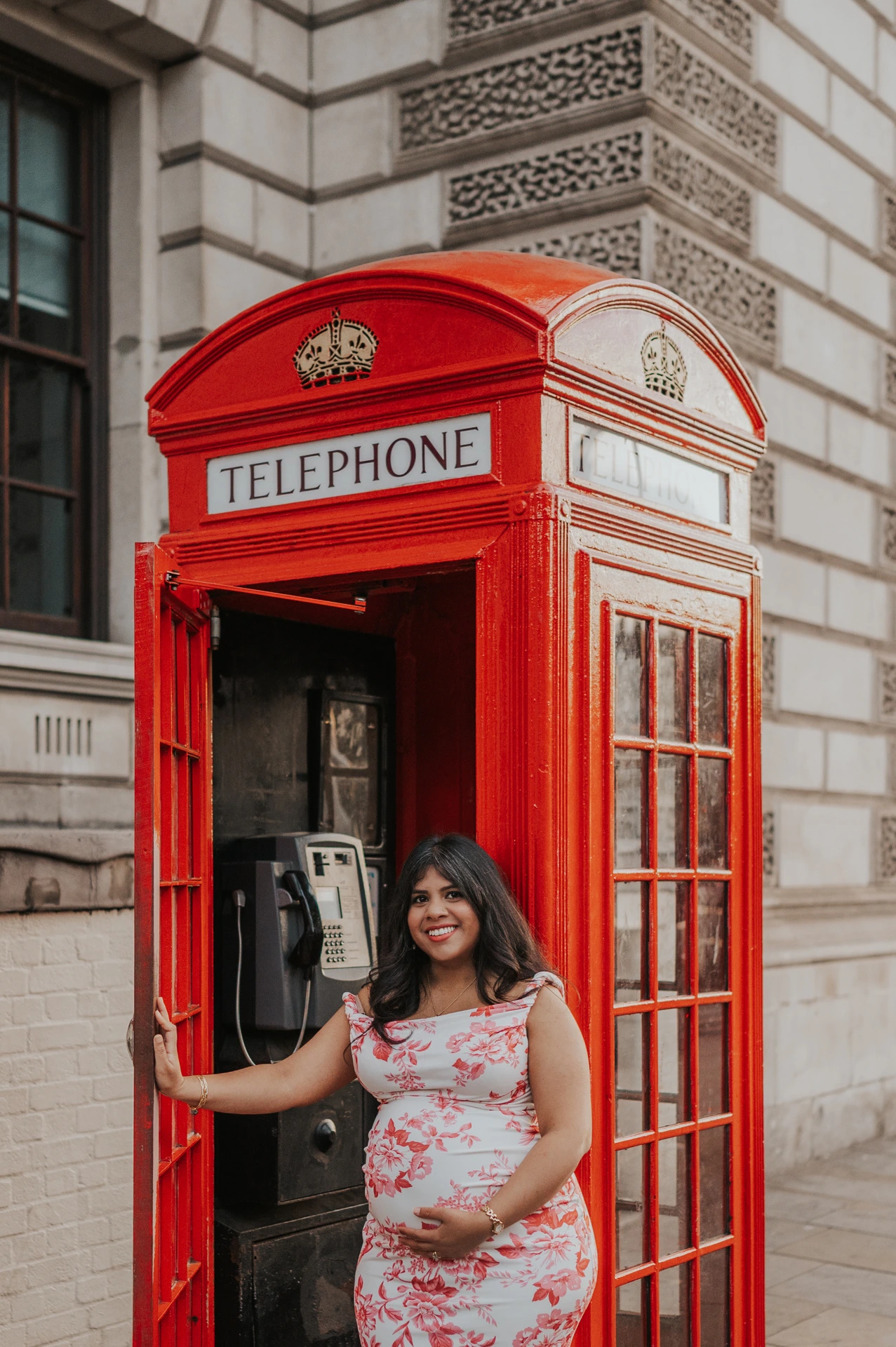 Closer portrait of the mother-to-be smiling at the open red telephone box door during her London maternity photoshoot.