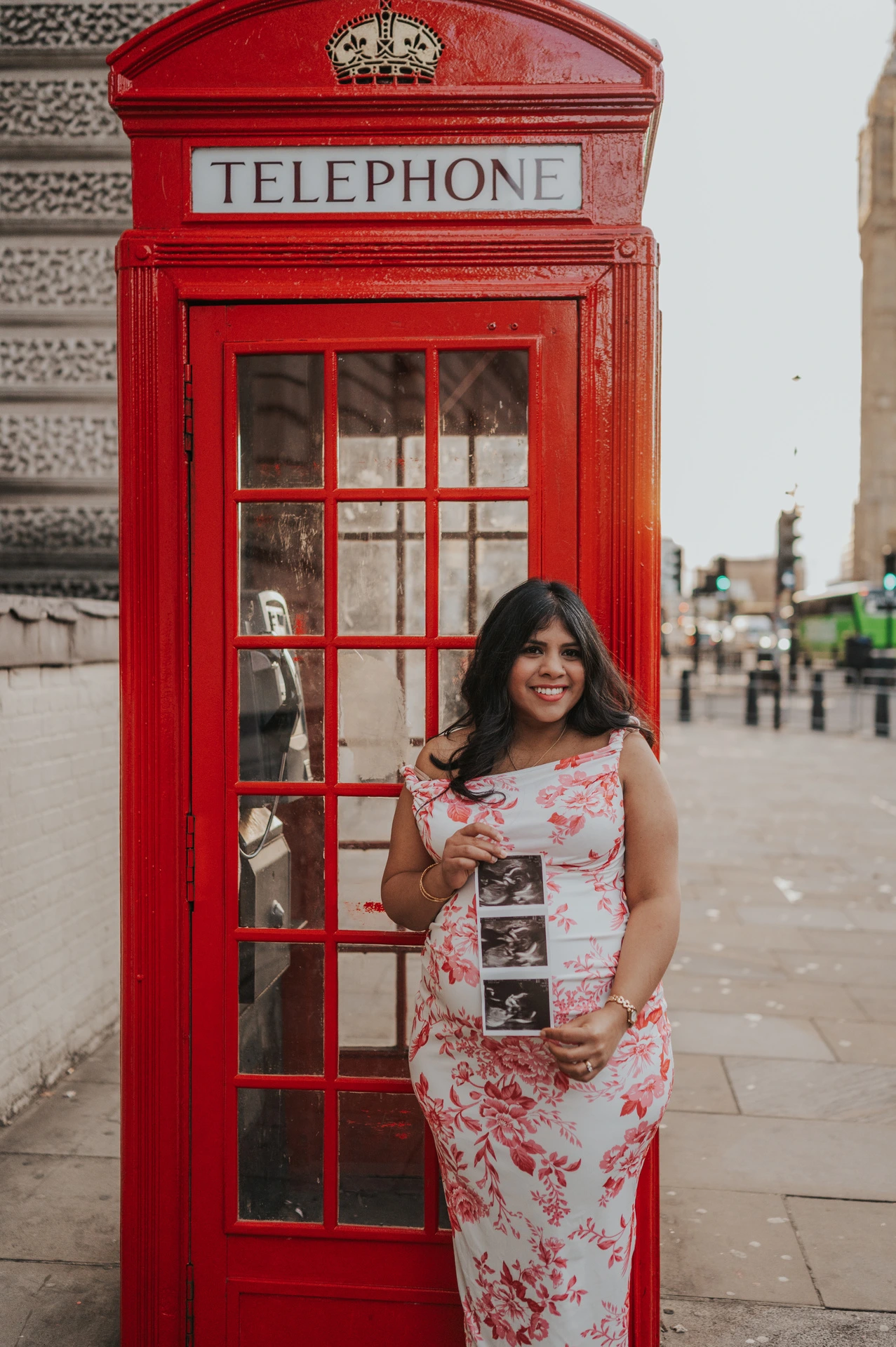 The mother-to-be smiling with an ultrasound scan held against her floral dress beside a red telephone box during her London maternity photoshoot.