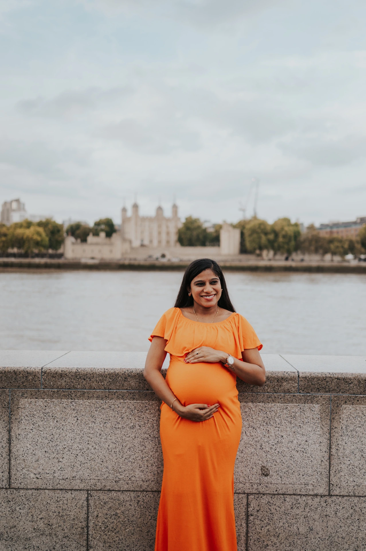 A pregnant woman in an orange dress stands beside the Queen’s Walk stone wall, with the Thames and Tower of London across the water during their London maternity photoshoot.