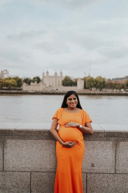 London maternity photoshoot: an orange-dress portrait facing the Tower of London. A pregnant woman in an orange dress stands beside the Queen’s Walk stone wall, with the Thames and Tower of London across the water during their London maternity photoshoot.