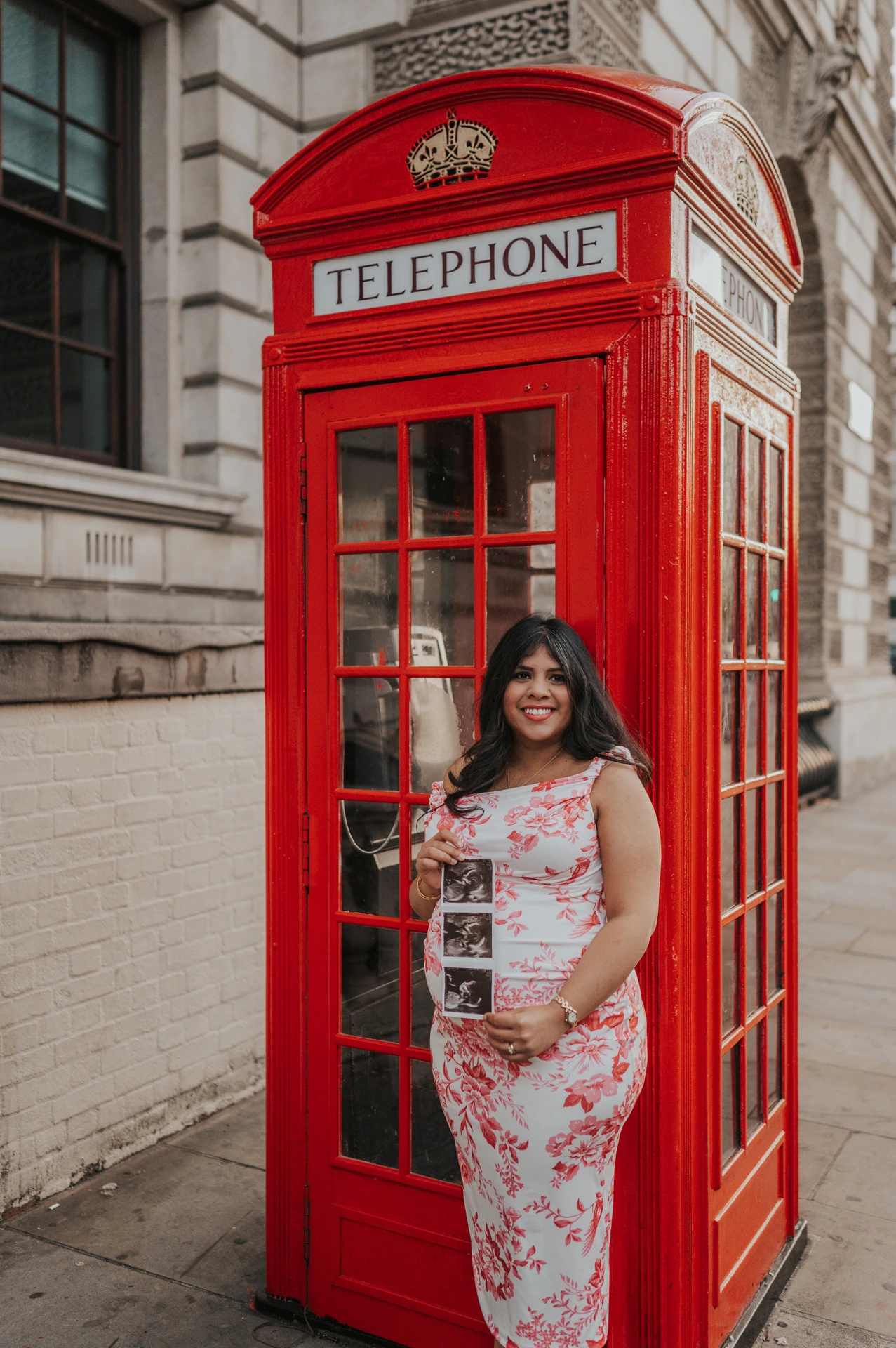The mother-to-be standing beside a red telephone box with her ultrasound scan and a view along the pavement during her London maternity photoshoot.