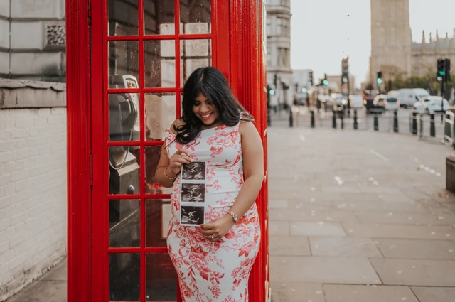 Landscape portrait of the mother-to-be looking down at her ultrasound scan in front of a red telephone box during her London maternity photoshoot.