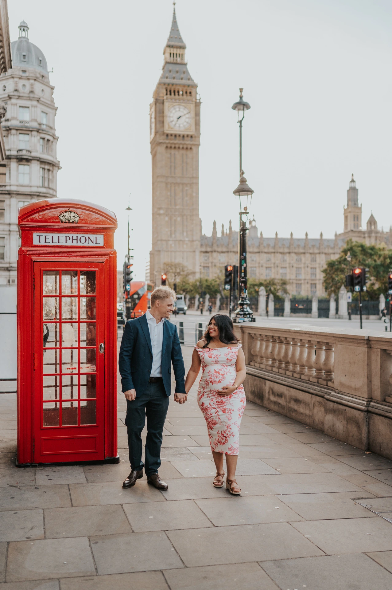 The expectant couple walking hand in hand past a red telephone box, with Big Ben in view, during their London maternity photoshoot.