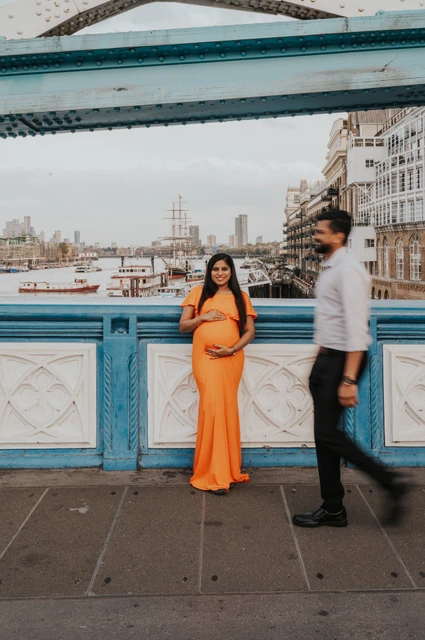 London maternity photoshoot: a moving portrait with Butler’s Wharf behind. A pregnant woman in an orange dress stands beside the blue Tower Bridge railing while her partner walks across the foreground, with Butler’s Wharf behind during their London maternity photoshoot.