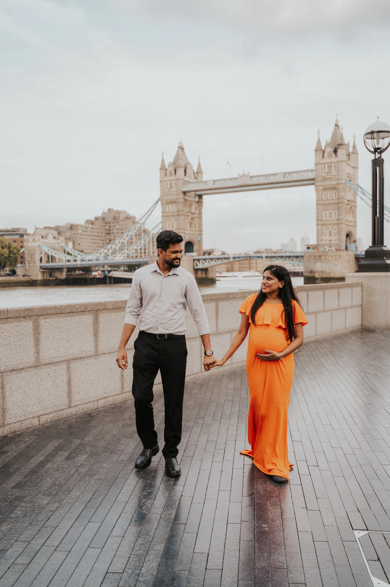 A couple walk hand in hand along the Queen’s Walk, with her orange dress beside the pale river wall and Tower Bridge behind during their London maternity photoshoot.