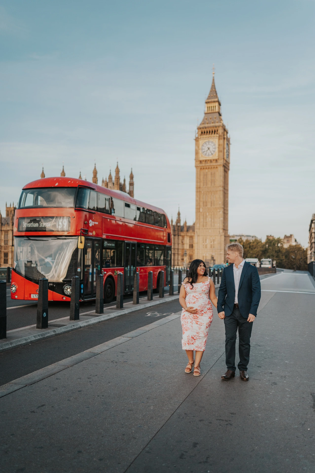 The couple walking on Westminster Bridge beside a passing red double-decker bus, with Big Ben behind, during their London maternity photoshoot.