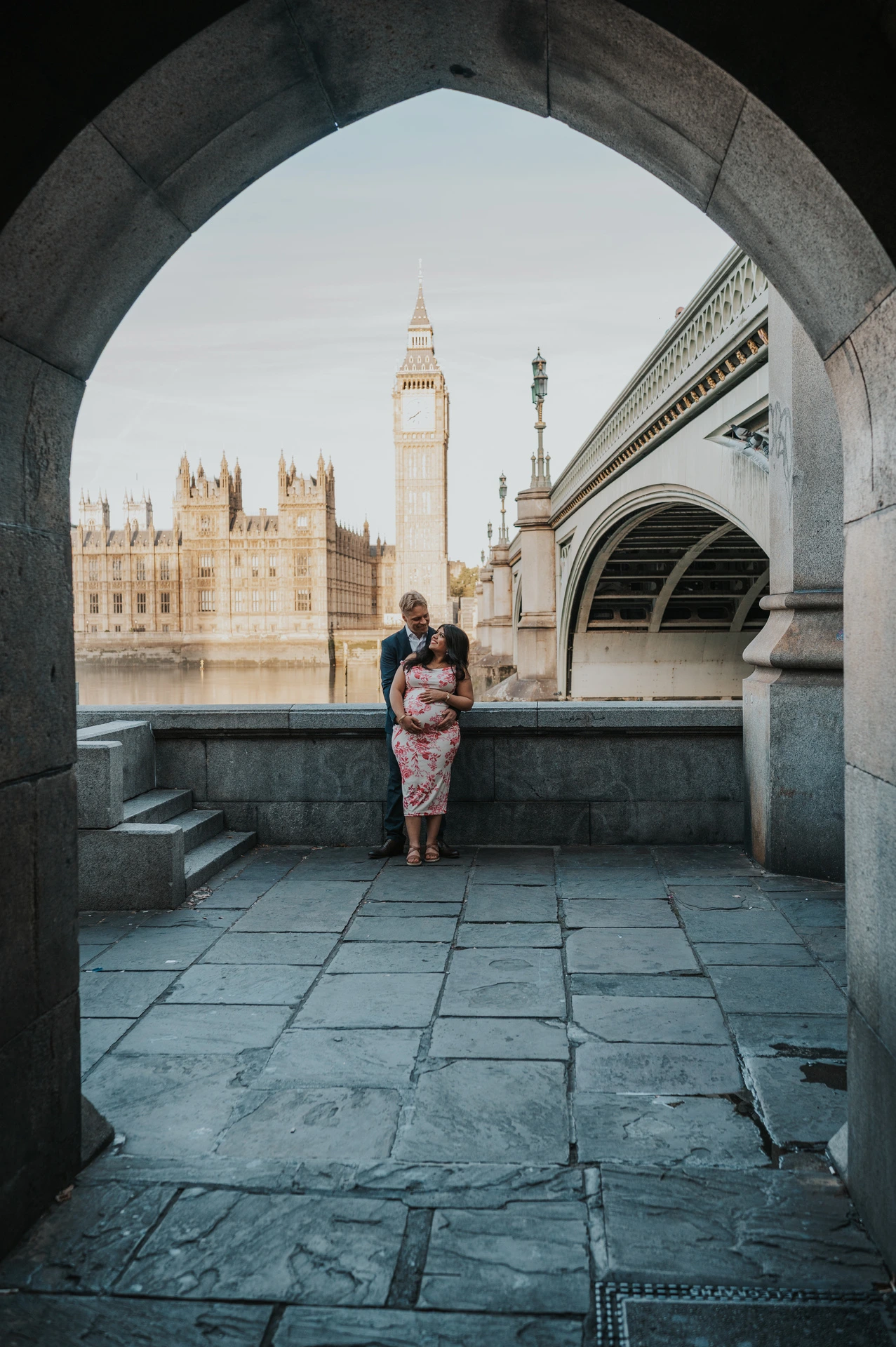 The couple framed by a stone arch beside Westminster Bridge, with Big Ben across the Thames, during their London maternity photoshoot.