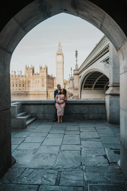 The couple framed by a stone arch beside Westminster Bridge, with Big Ben across the Thames, during their London maternity photoshoot.