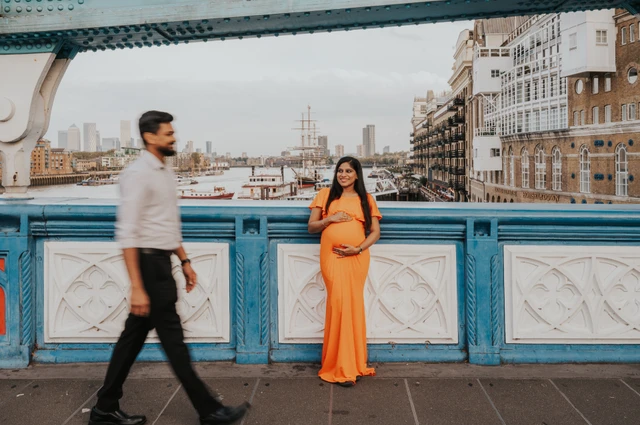 A pregnant woman in an orange dress stands beside the blue-and-white Tower Bridge railing as her partner walks across the wide foreground, with Butler’s Wharf beyond during their London maternity photoshoot.
