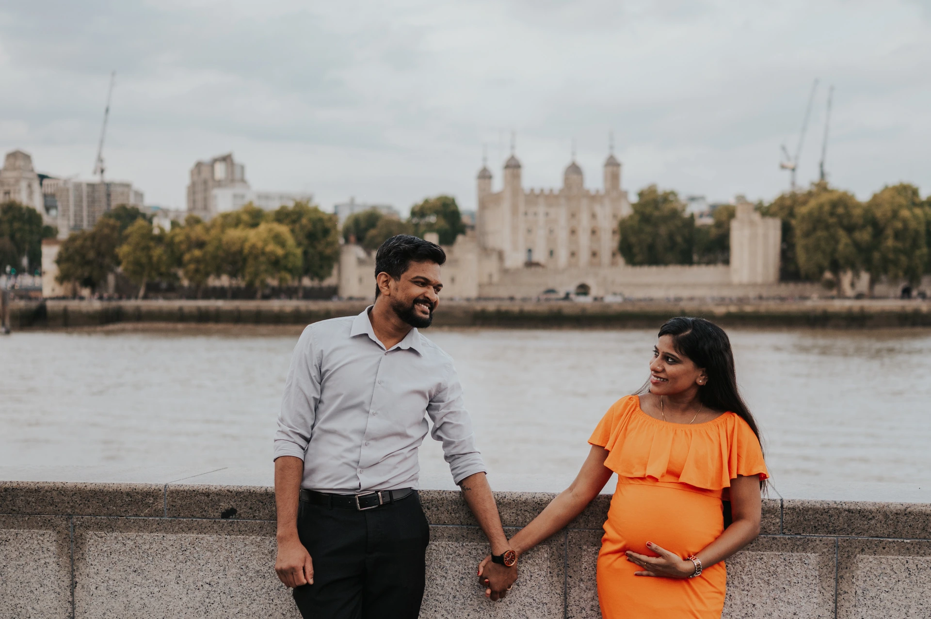 A wide portrait shows a couple smiling towards each other while holding hands at the river wall, with the Tower of London across the Thames during their London maternity photoshoot.