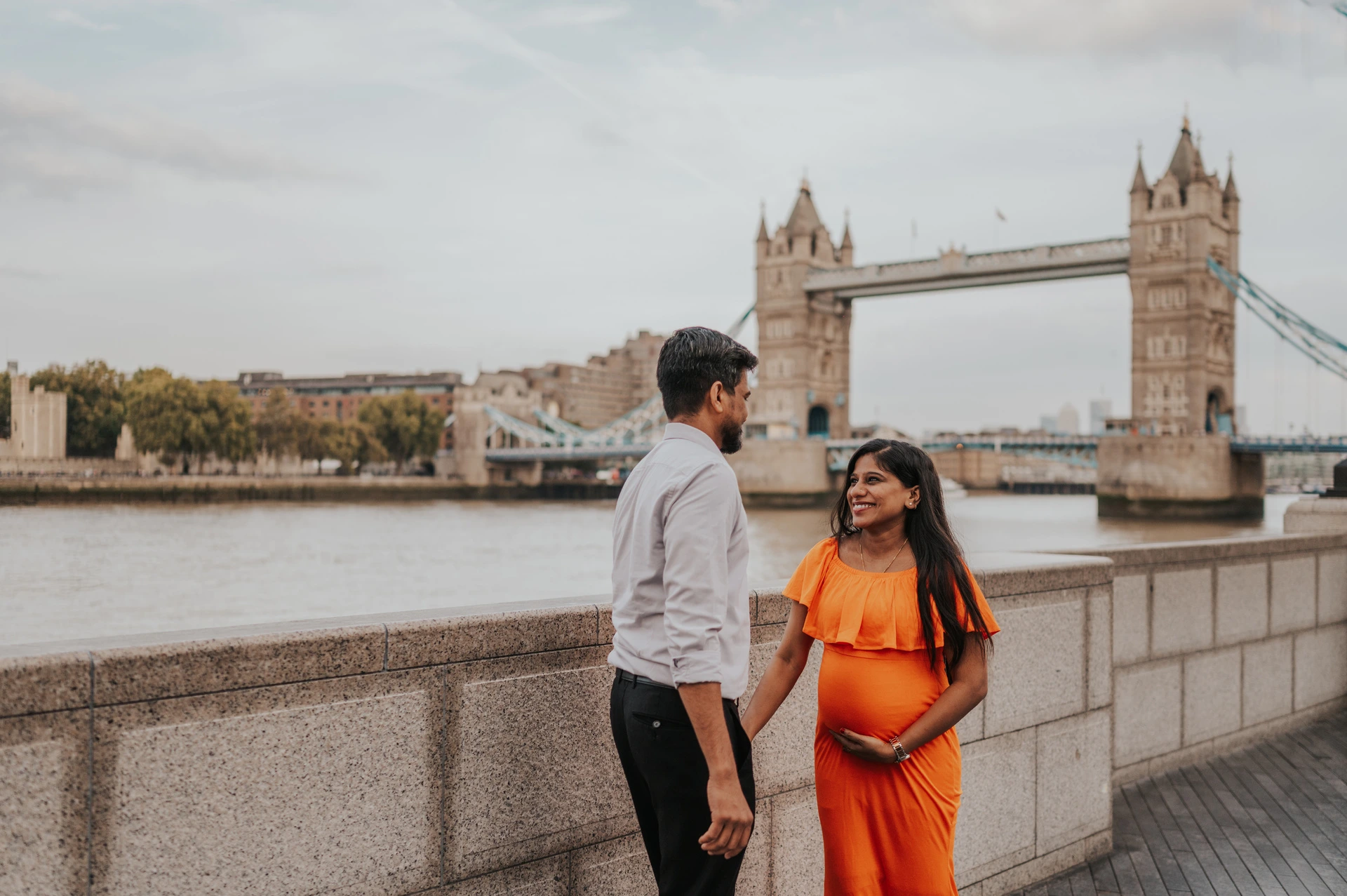 London maternity photoshoot: a wide couple portrait with Tower Bridge. A wide portrait shows a couple beside the Queen’s Walk river wall, with her orange dress in the foreground and the full span of Tower Bridge beyond during their London maternity photoshoot.